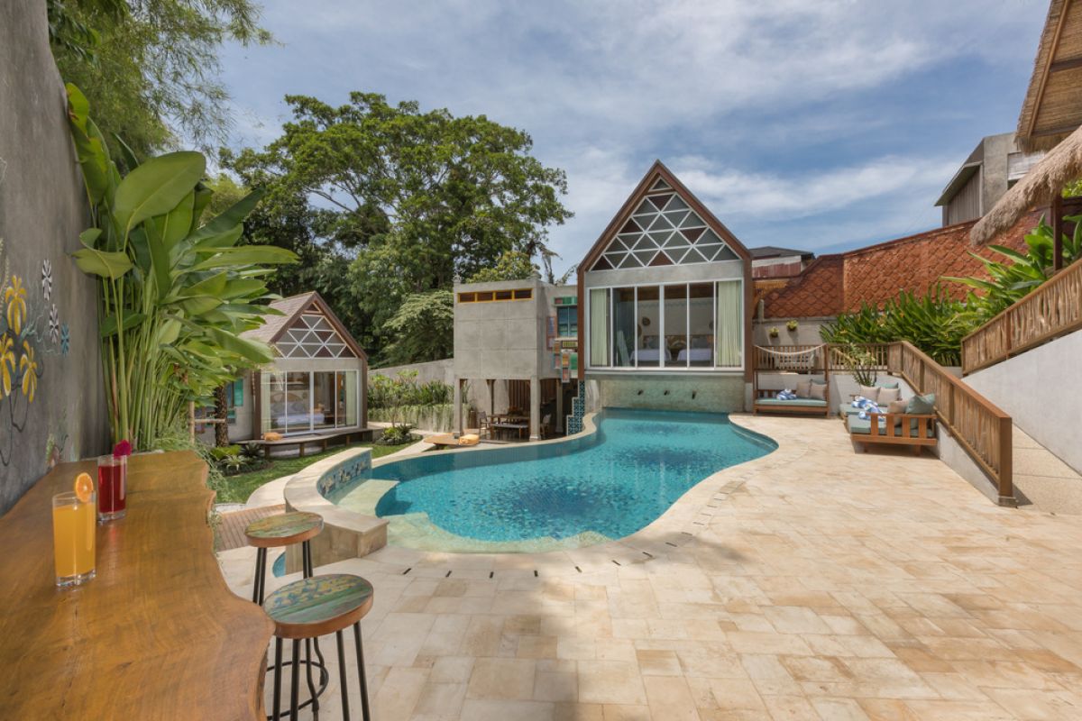 Spacious tropical Balinese villa interior with high wooden-beamed ceiling, large glass windows overlooking lush garden, plush sectional sofa, and modern kitchen bar. - Bali Villas