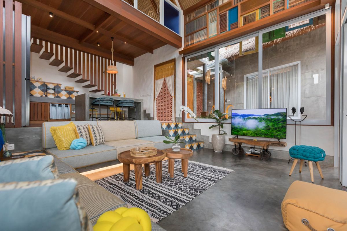 Spacious tropical Balinese villa interior with high wooden-beamed ceiling, large glass windows overlooking lush garden, plush sectional sofa, and modern kitchen bar. - Bali Villas