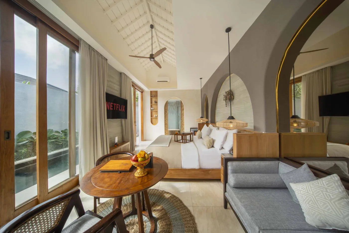 Spacious luxury tropical bedroom with king bed, wooden accents, ceiling fan, and poolside garden view through large windows. - Bali Villas