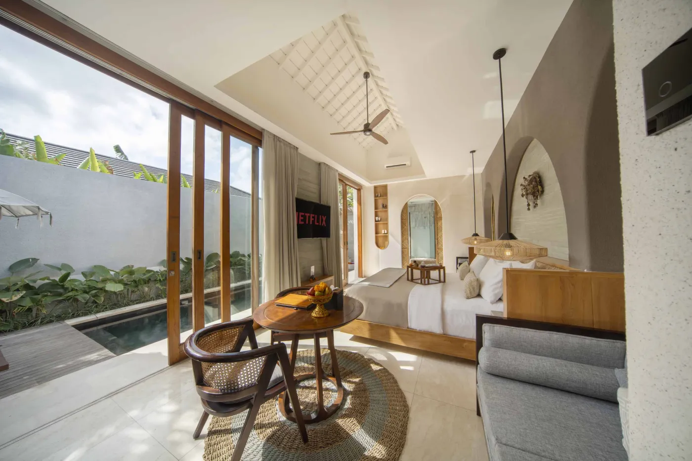 Spacious luxury tropical bedroom with king bed, wooden accents, ceiling fan, and poolside garden view through large windows. - Bali Villas