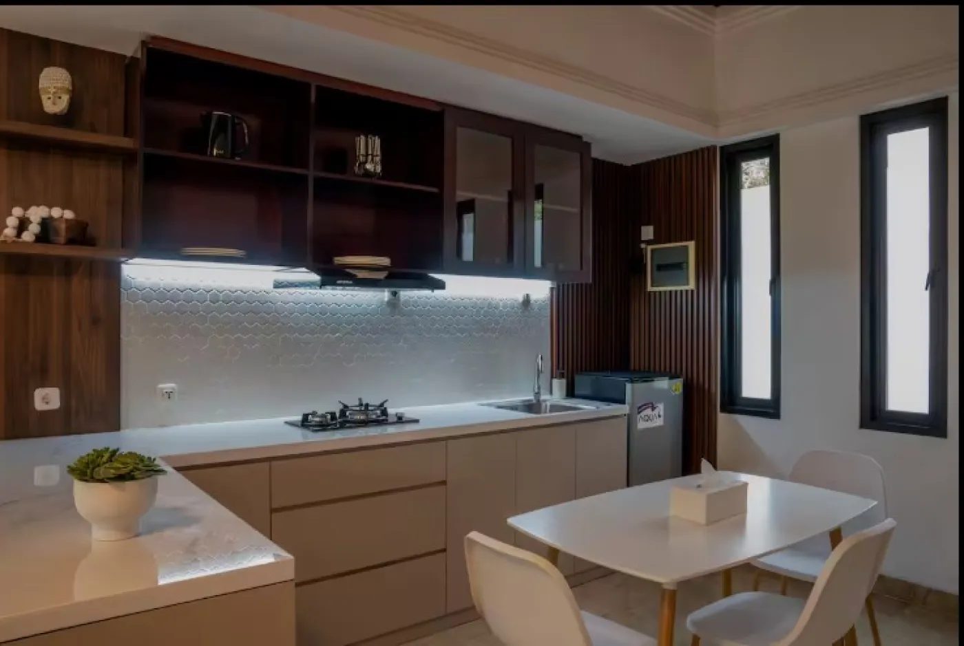 Modern wooden kitchen interior with white countertops, gas stove, sink, LED-lit backsplash, open shelves, and a small dining table with chairs. - Bali Villas