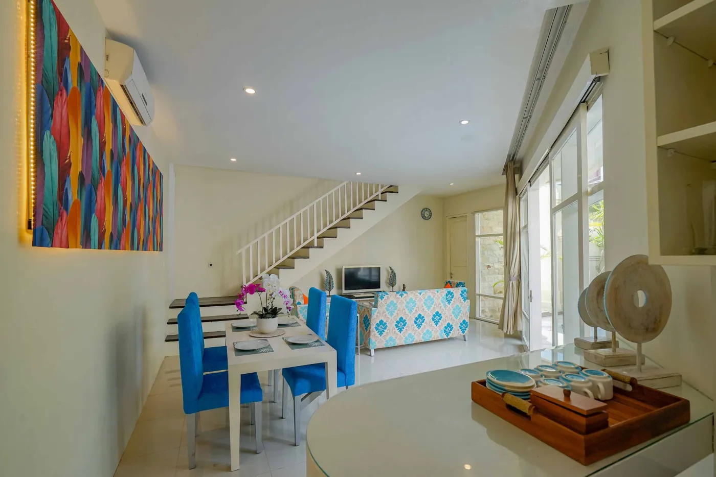 Bright modern tropical villa interior with colorful feather wall art, wooden dining table in blue chairs, and adjacent living area with patterned sofa. - Bali Villas