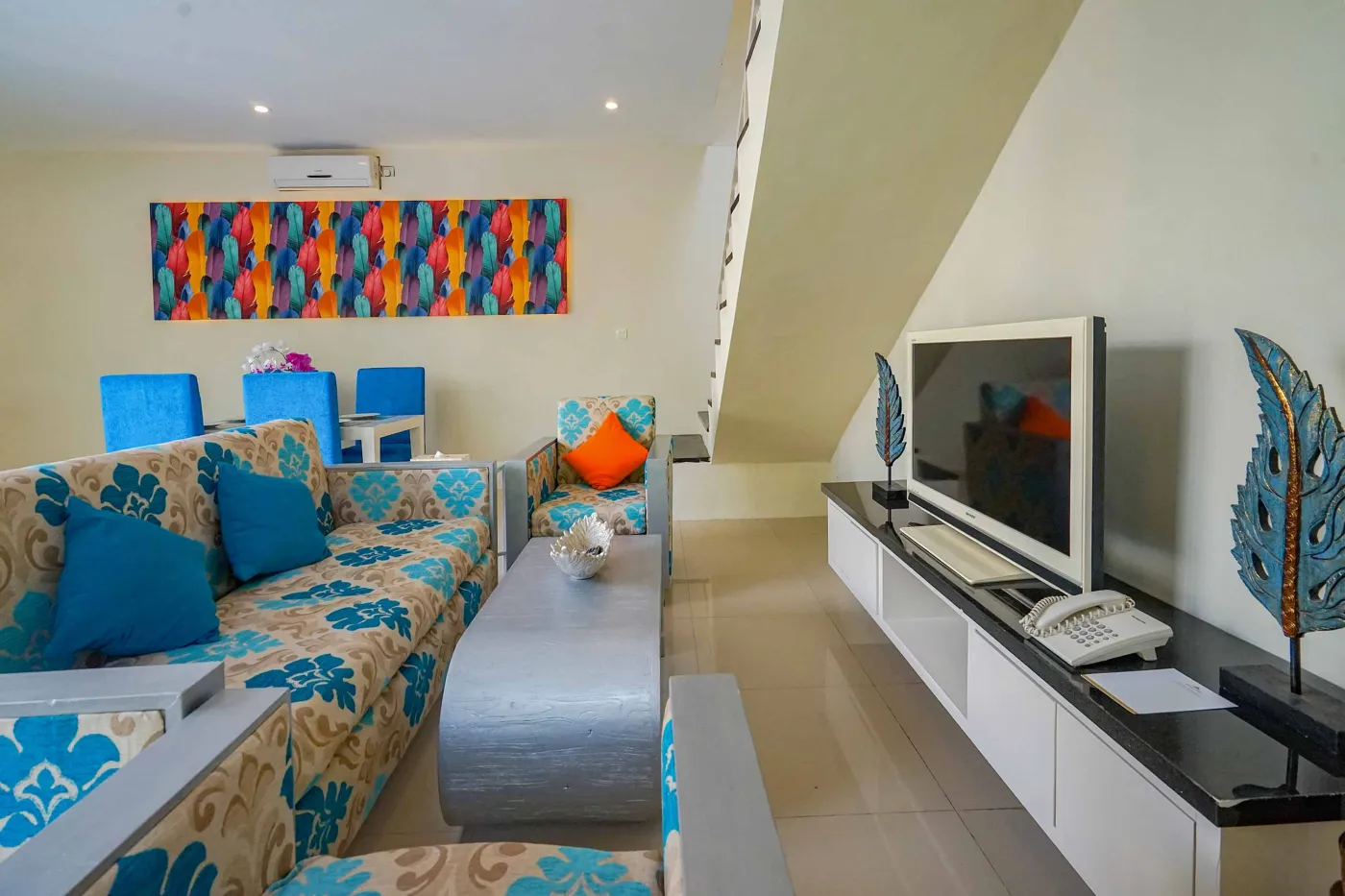 Bright modern tropical villa interior with colorful feather wall art, wooden dining table in blue chairs, and adjacent living area with patterned sofa. - Bali Villas