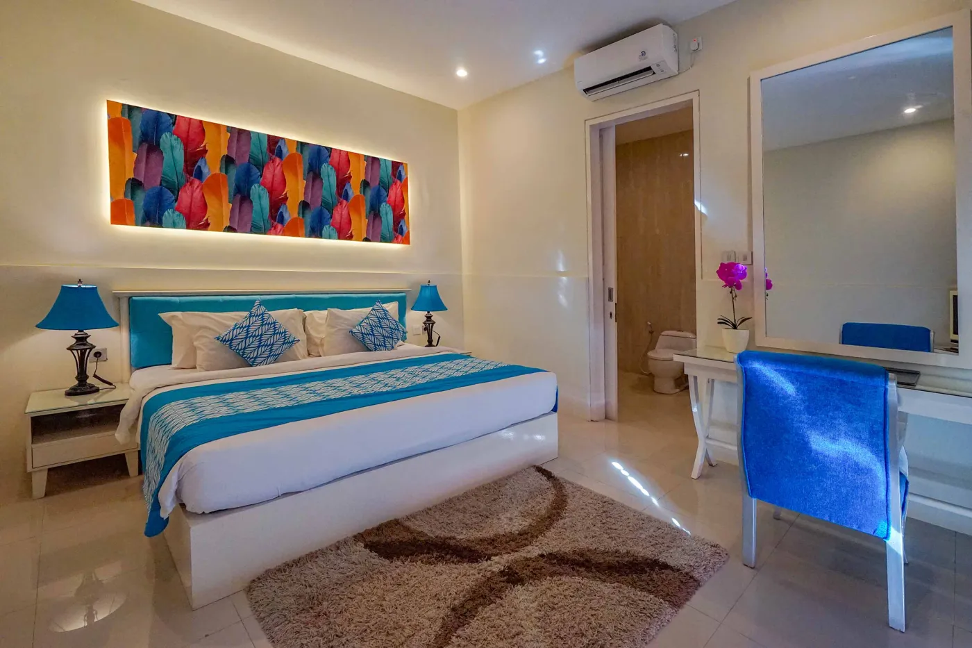 Bright modern tropical villa interior with colorful feather wall art, wooden dining table in blue chairs, and adjacent living area with patterned sofa. - Bali Villas