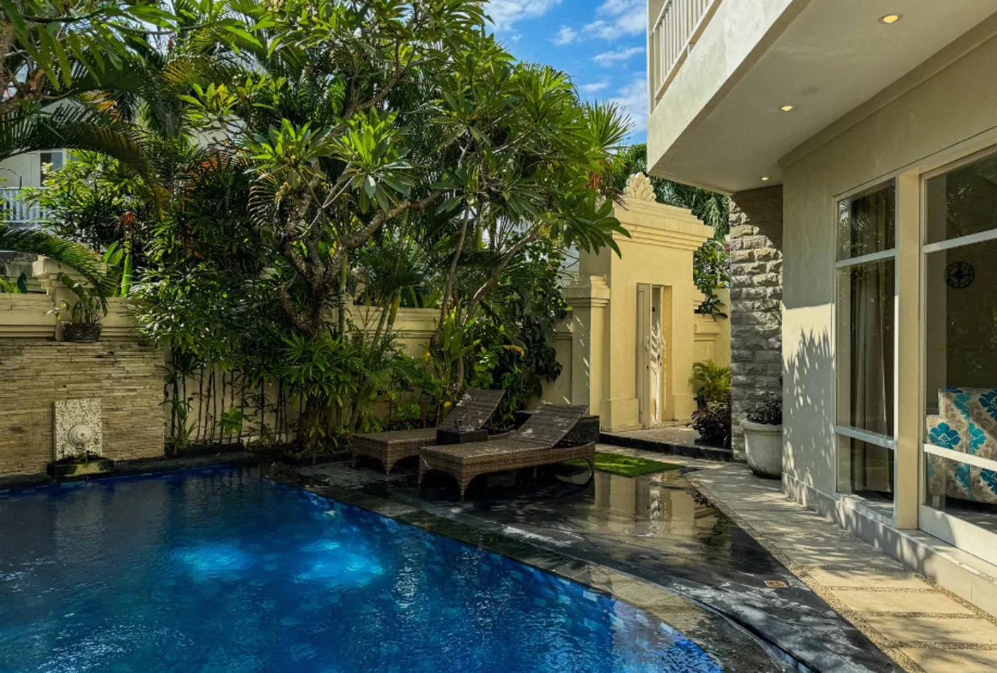 Bright modern tropical villa interior with colorful feather wall art, wooden dining table in blue chairs, and adjacent living area with patterned sofa. - Bali Villas