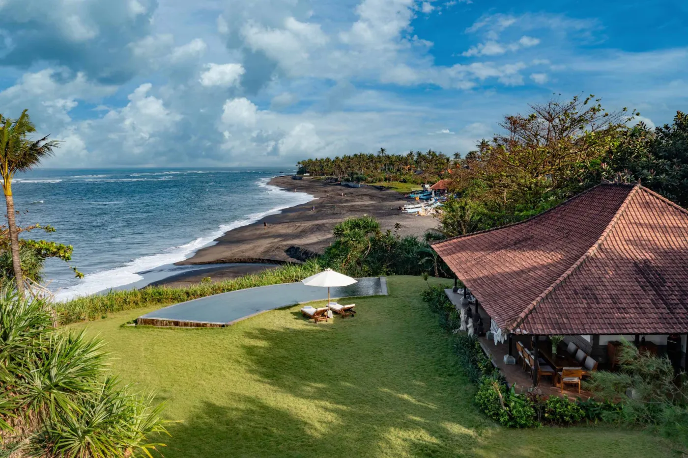 Luxurious Balinese veranda at sunset overlooking ocean and infinity pool, with wooden furniture, tropical plants, and cushioned seating. - Bali Villas