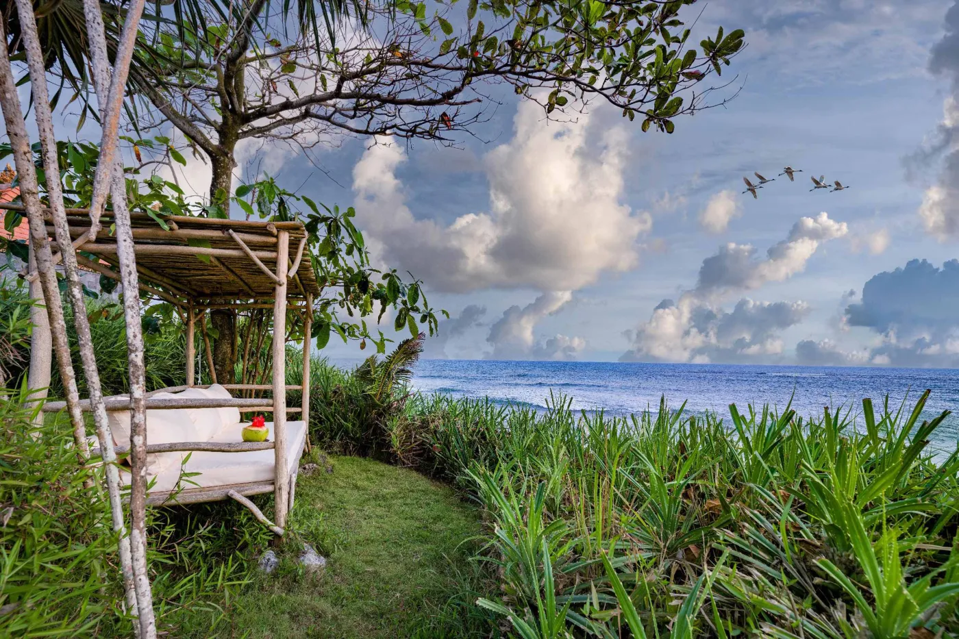 Luxurious Balinese veranda at sunset overlooking ocean and infinity pool, with wooden furniture, tropical plants, and cushioned seating. - Bali Villas