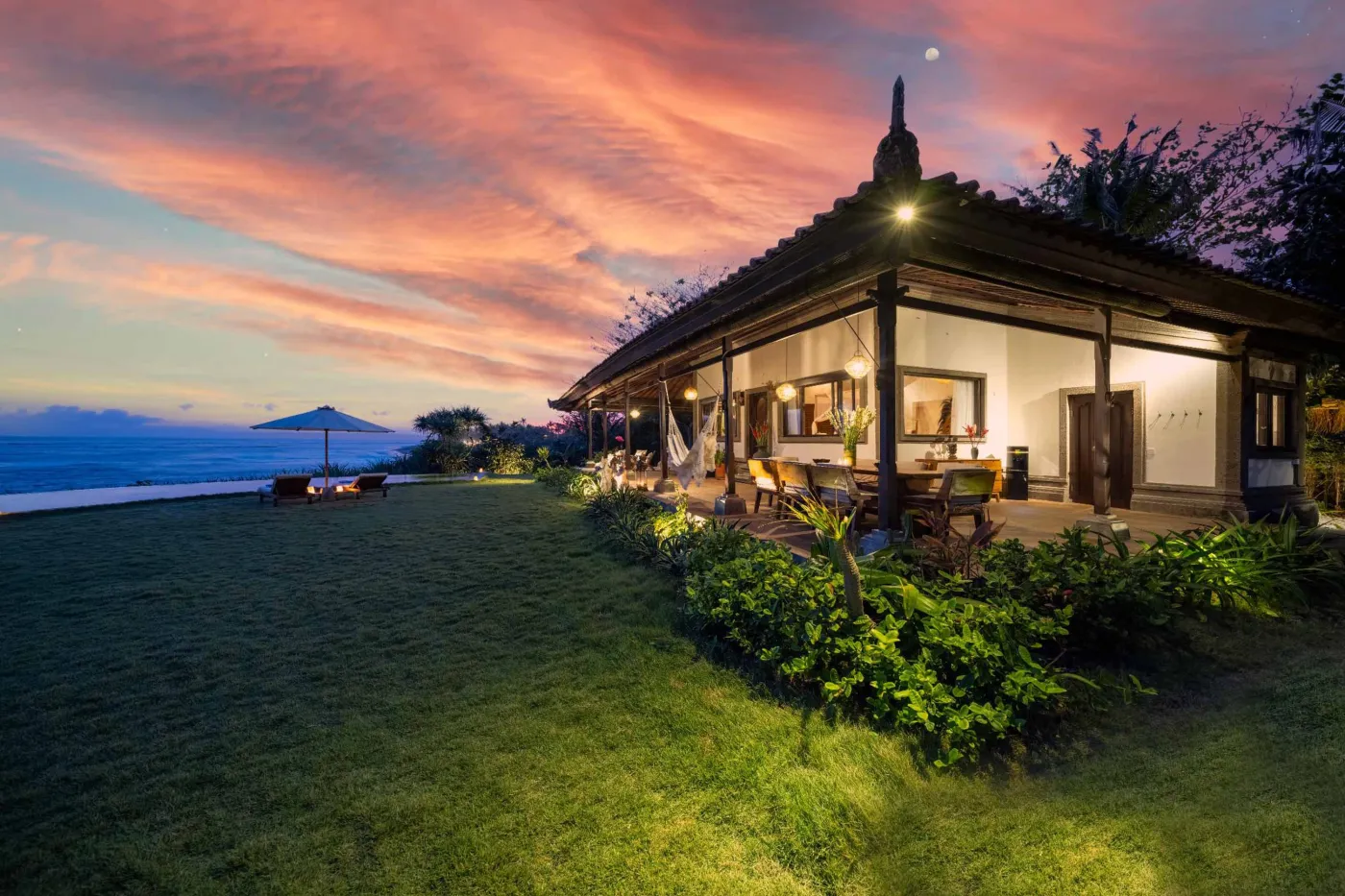 Luxurious Balinese veranda at sunset overlooking ocean and infinity pool, with wooden furniture, tropical plants, and cushioned seating. - Bali Villas