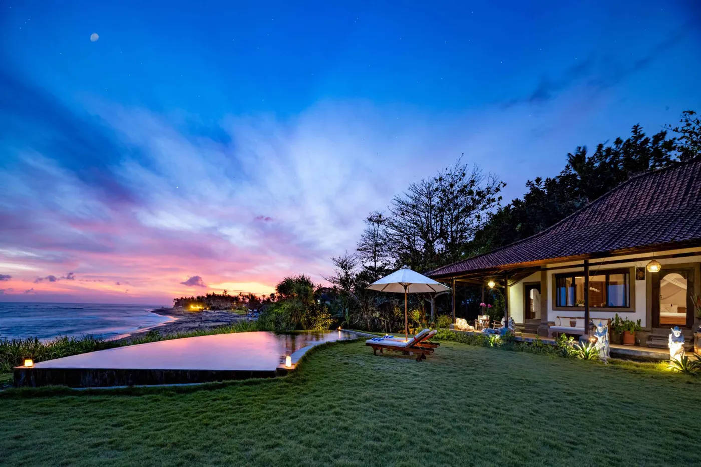 Luxurious Balinese veranda at sunset overlooking ocean and infinity pool, with wooden furniture, tropical plants, and cushioned seating. - Bali Villas