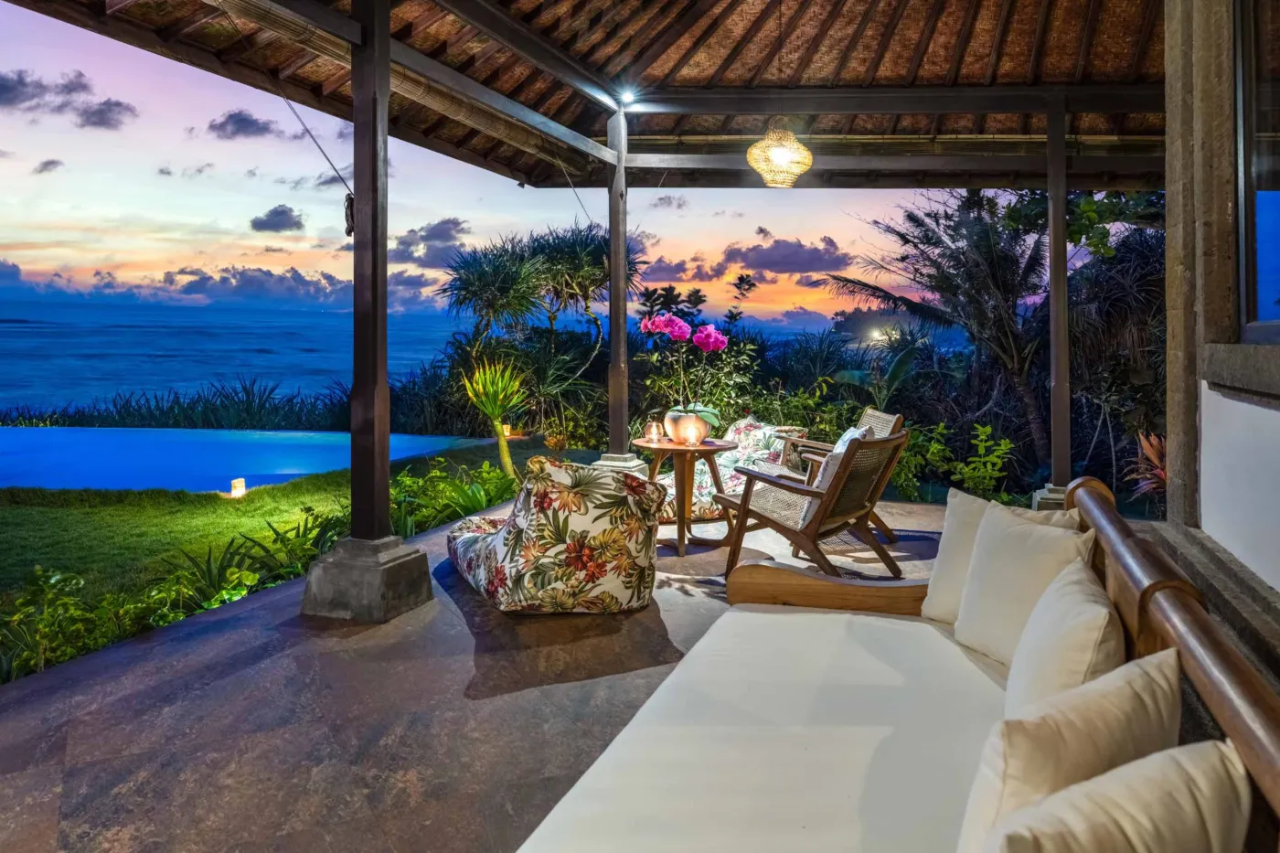 Luxurious Balinese veranda at sunset overlooking ocean and infinity pool, with wooden furniture, tropical plants, and cushioned seating. - Bali Villas