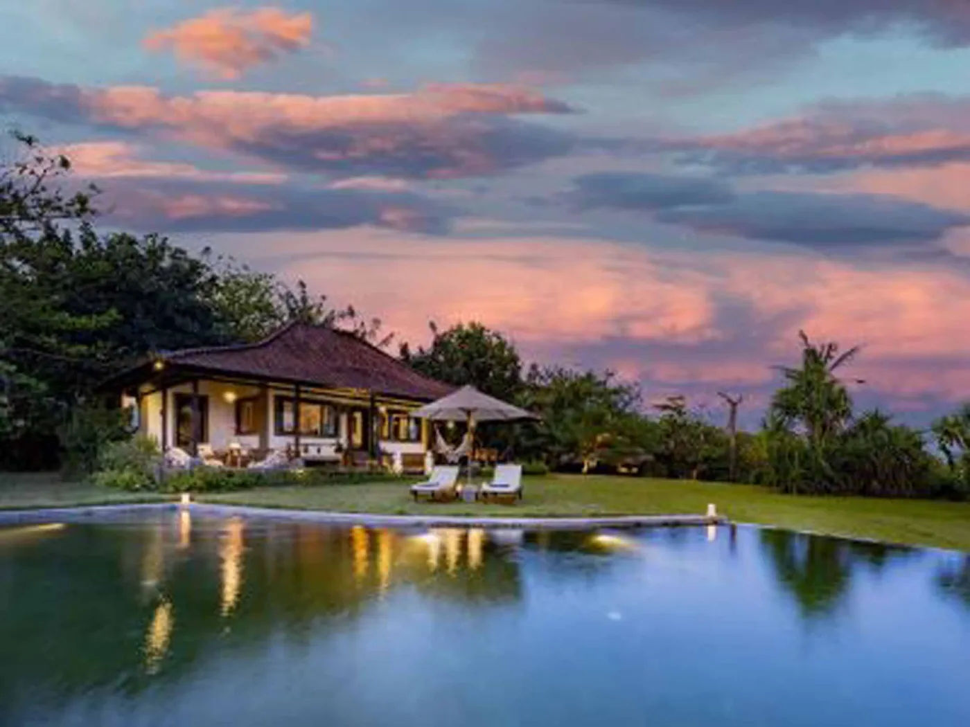 Luxurious Balinese veranda at sunset overlooking ocean and infinity pool, with wooden furniture, tropical plants, and cushioned seating. - Bali Villas