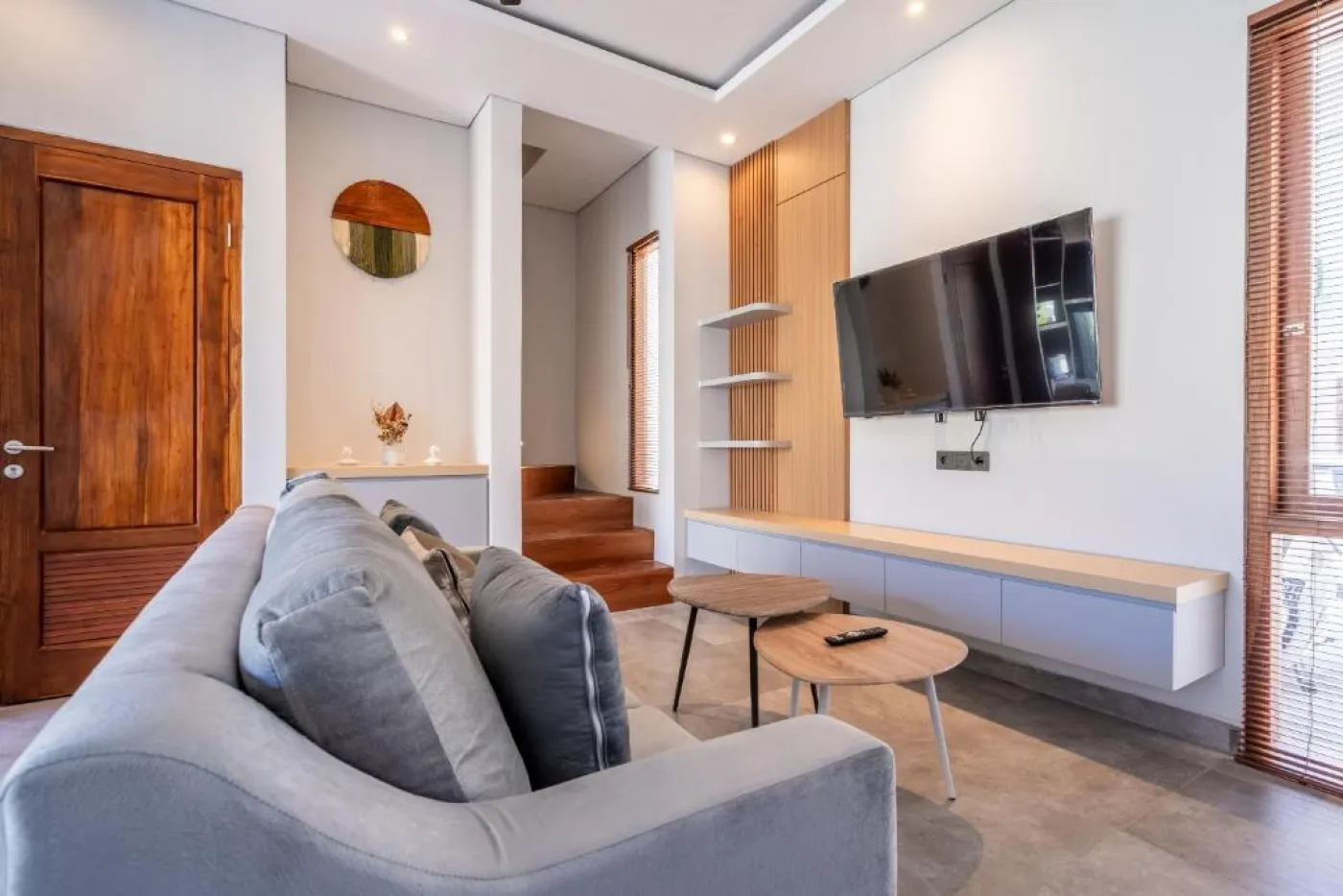 Modern Balinese-style bathroom with glass shower enclosure, round wooden mirror, stone vanity, and stacked towels. - Bali Villas