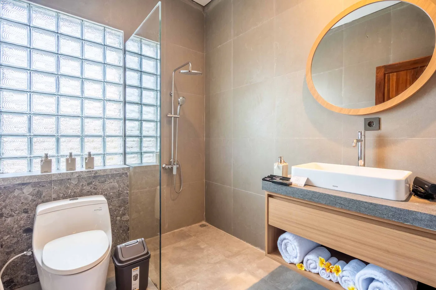 Modern Balinese-style bathroom with glass shower enclosure, round wooden mirror, stone vanity, and stacked towels. - Bali Villas