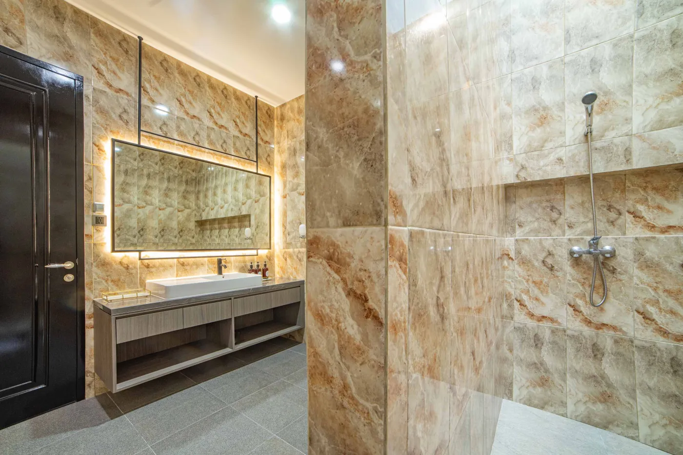 Modern luxury bathroom with LED-lit mirror, wooden vanity sink, white toilet, and beige marble-patterned tiled walls. - Bali Villas