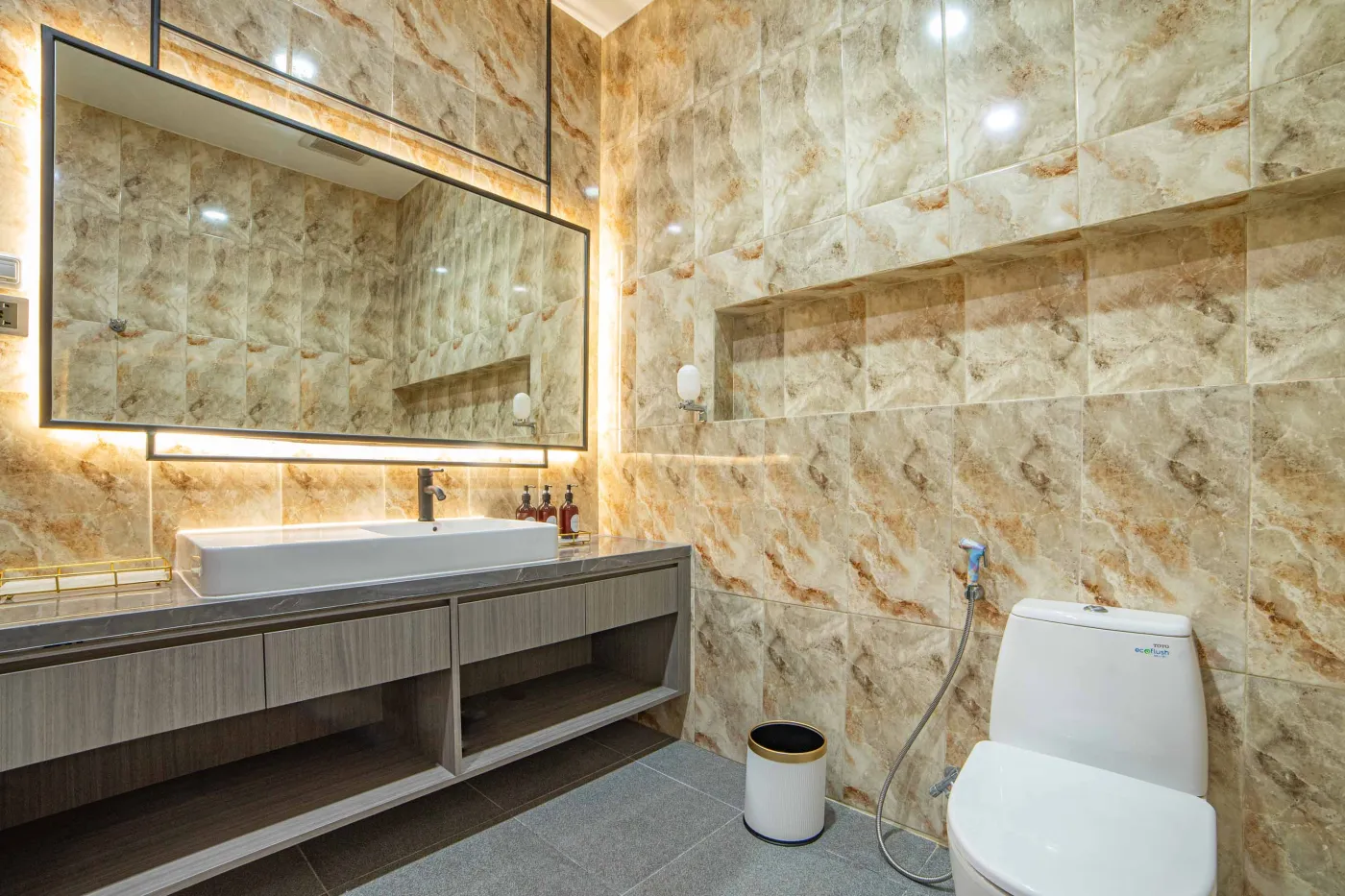 Modern luxury bathroom with LED-lit mirror, wooden vanity sink, white toilet, and beige marble-patterned tiled walls. - Bali Villas