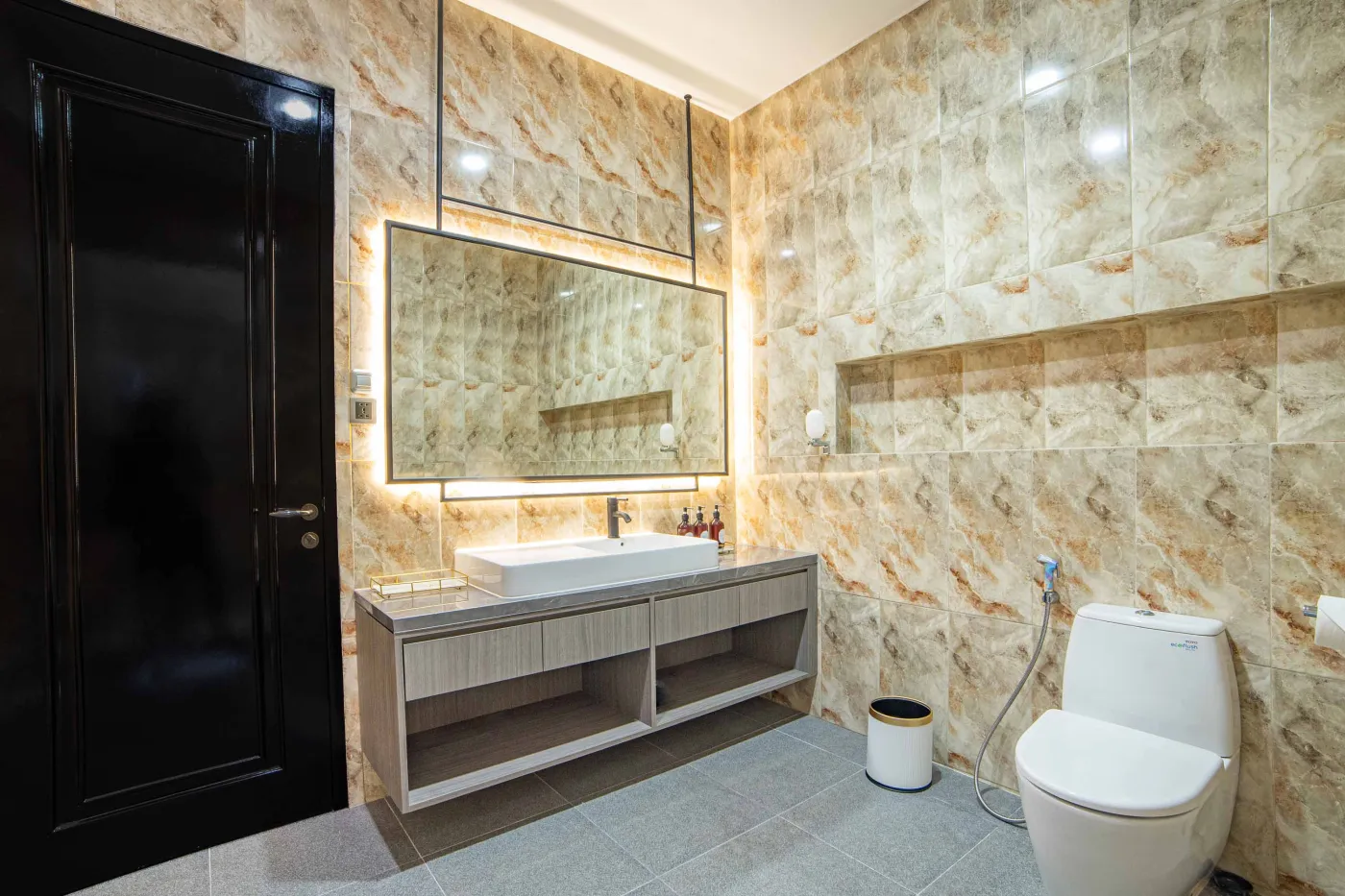 Modern luxury bathroom with LED-lit mirror, wooden vanity sink, white toilet, and beige marble-patterned tiled walls. - Bali Villas