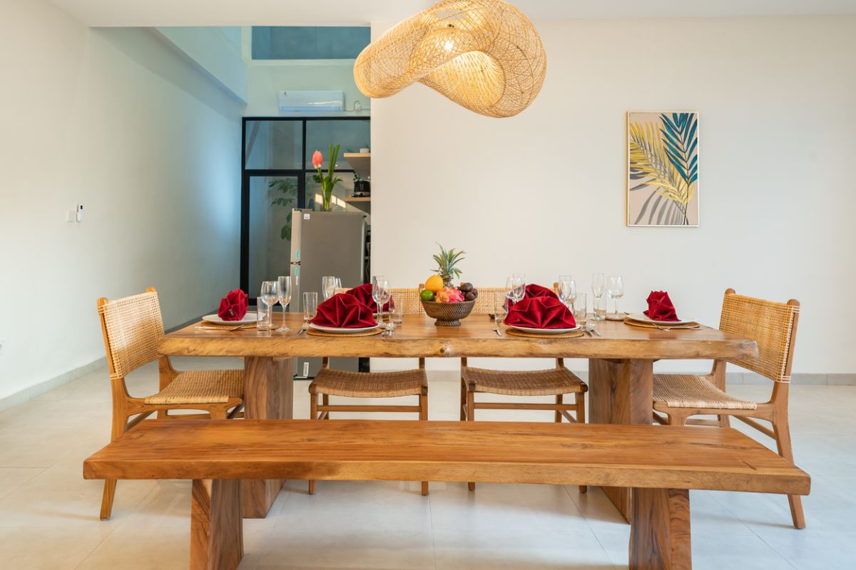Modern tropical dining room with rustic wooden table, benches, red napkins, fruit bowls, and woven hanging lamp. - Bali Villas