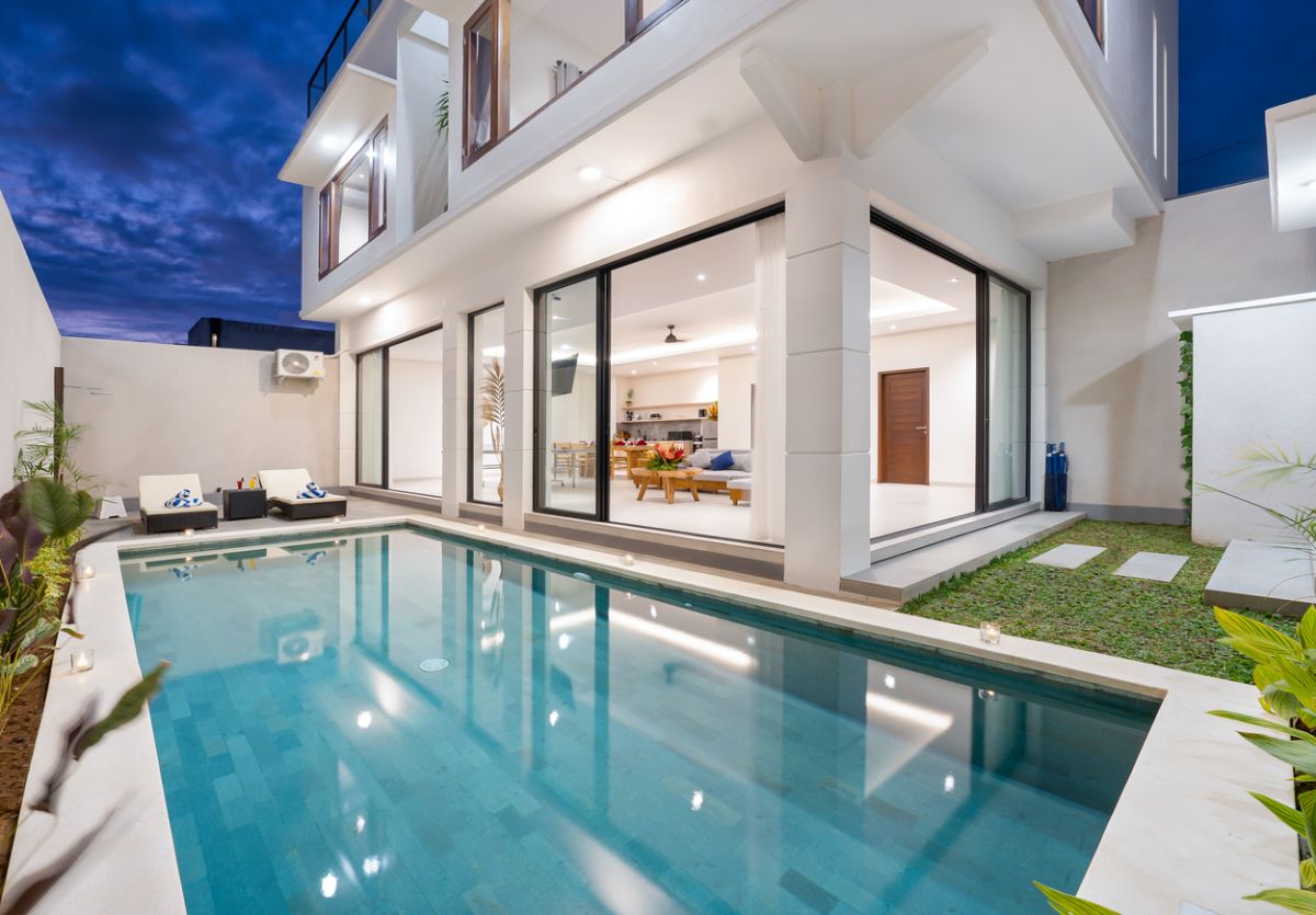 Modern luxury villa with turquoise infinity pool, lounge chairs, and open glass living area at dusk in Bali. - Bali Villas
