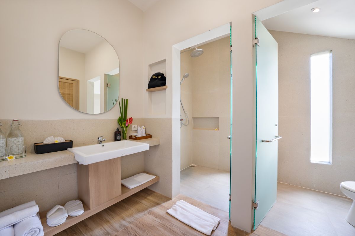 Modern minimalist bathroom interior with light wood vanity, oval mirror, tropical plants, open glass shower enclosure, and neutral tones. - Bali Villas