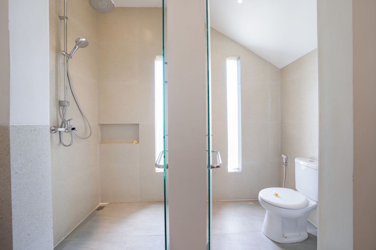 Modern minimalist bathroom interior with light wood vanity, oval mirror, tropical plants, open glass shower enclosure, and neutral tones. - Bali Villas