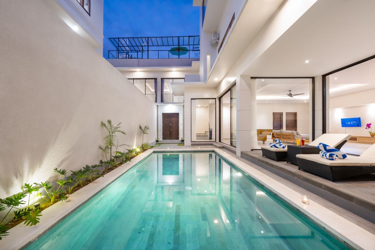 Modern tropical kitchen and dining area with wooden table, rattan chairs, hanging lamps, and infinity pool view through glass doors. - Bali Villas