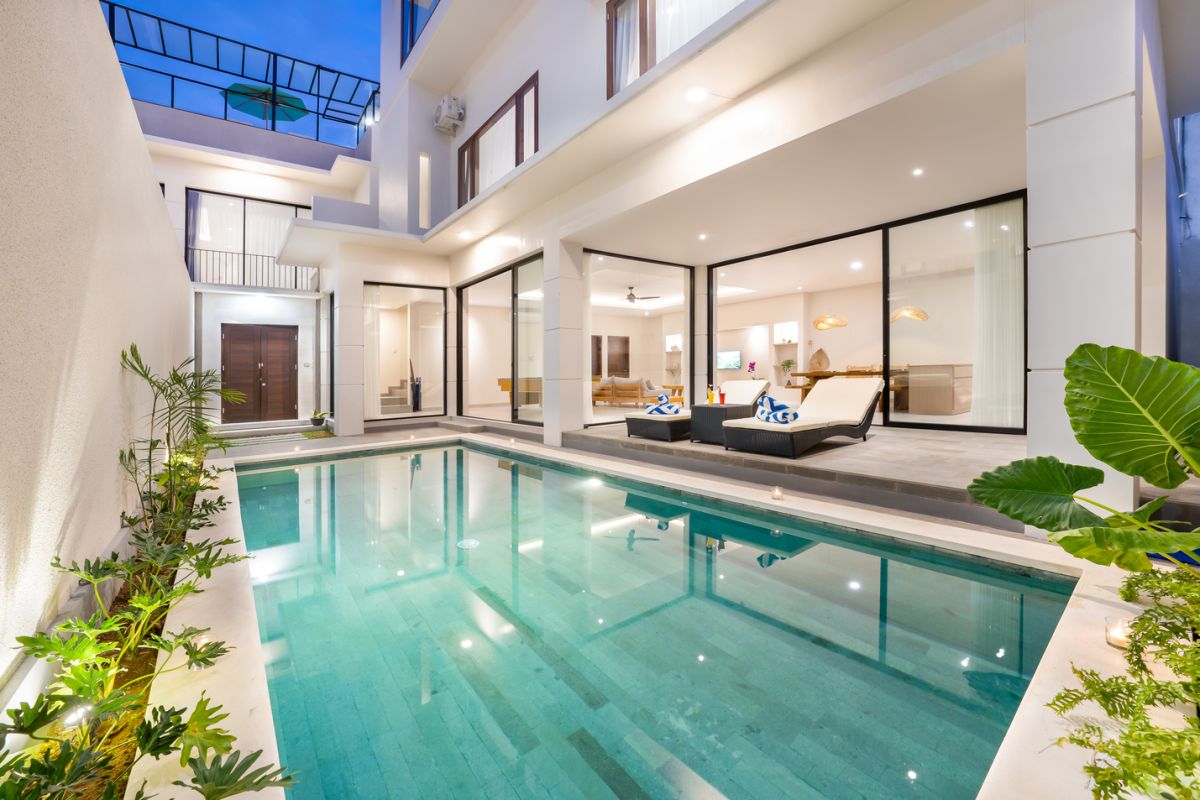 Modern tropical kitchen and dining area with wooden table, rattan chairs, hanging lamps, and infinity pool view through glass doors. - Bali Villas
