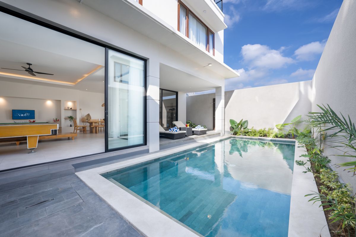 Modern tropical kitchen and dining area with wooden table, rattan chairs, hanging lamps, and infinity pool view through glass doors. - Bali Villas
