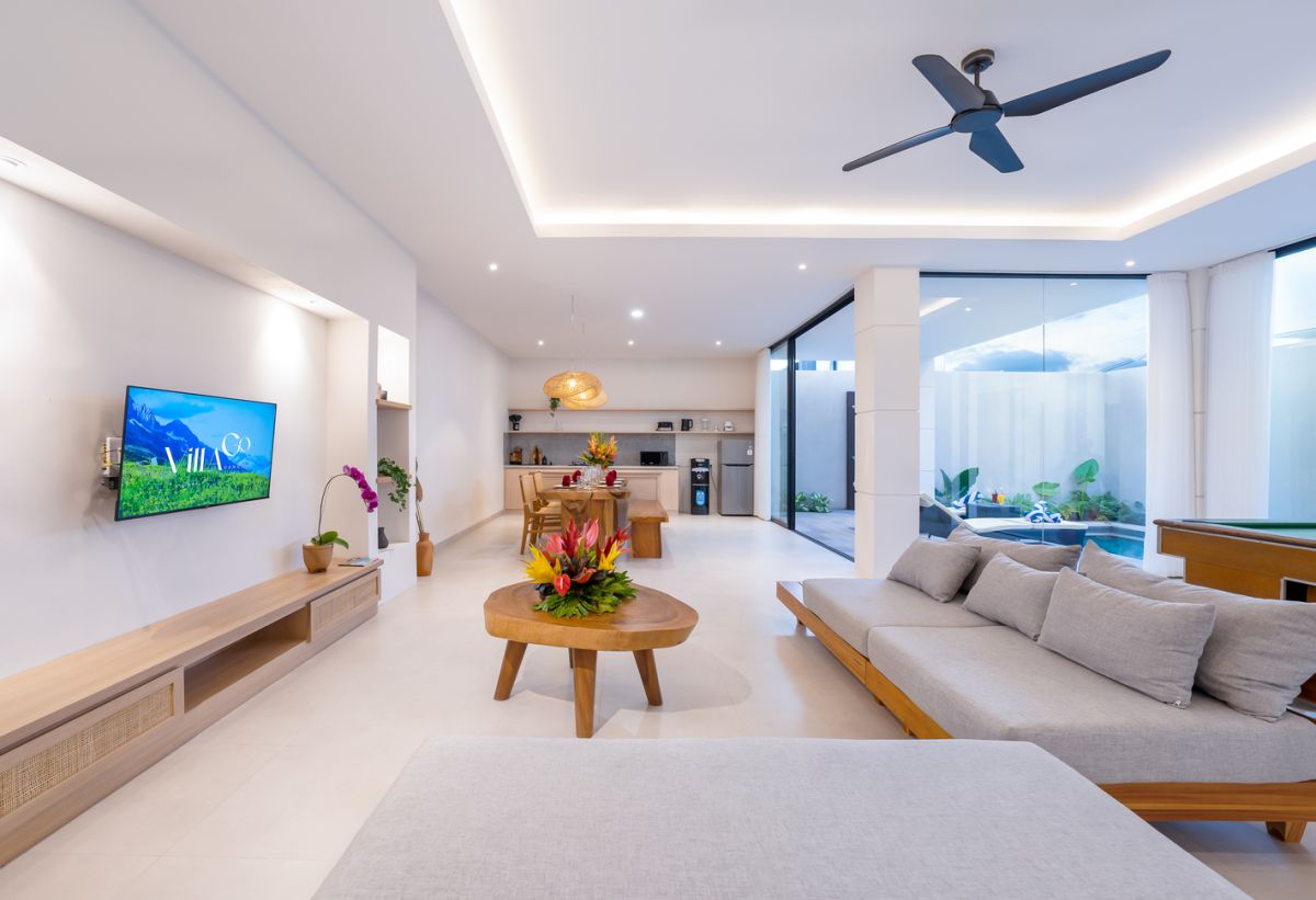 Modern tropical kitchen and dining area with wooden table, rattan chairs, hanging lamps, and infinity pool view through glass doors. - Bali Villas