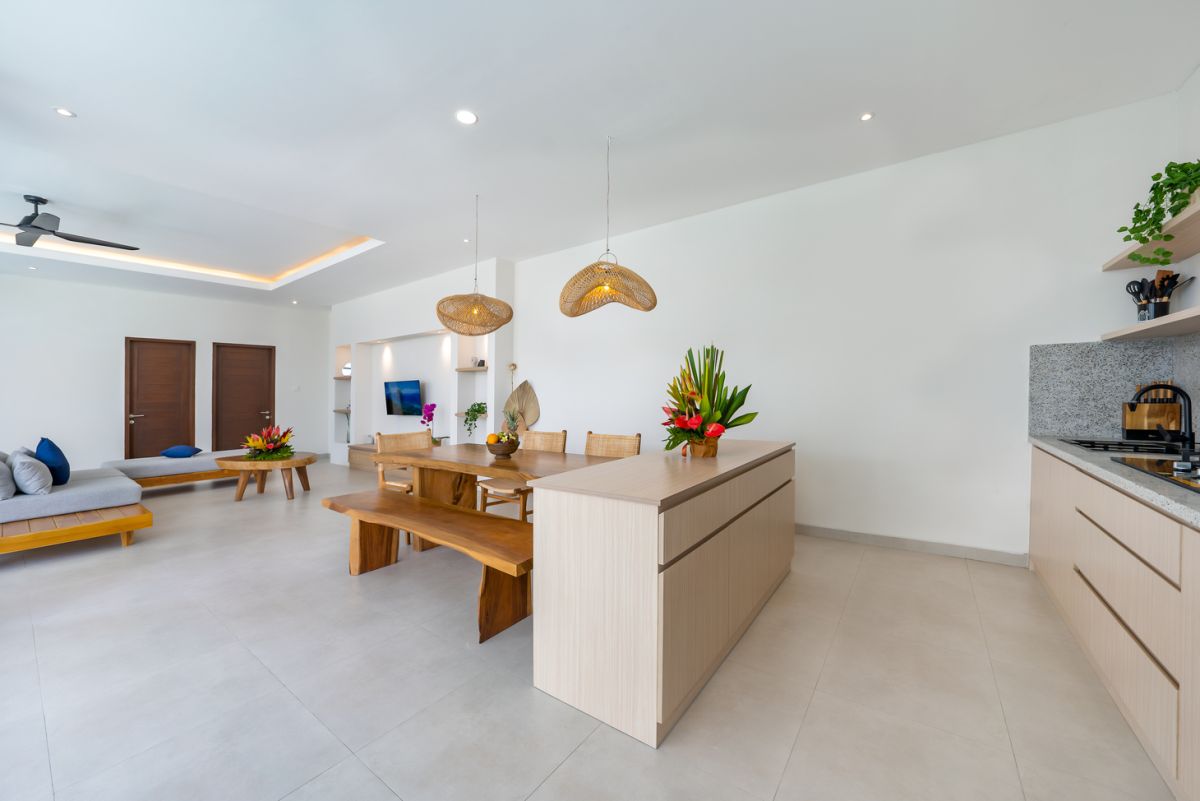 Modern tropical kitchen and dining area with wooden table, rattan chairs, hanging lamps, and infinity pool view through glass doors. - Bali Villas