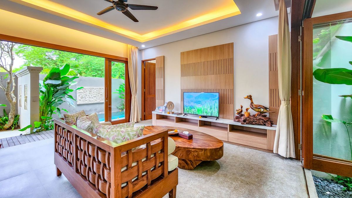 Tropical Balinese outdoor lounge with blue cushions on wooden deck, bamboo pergola, ceiling fan, lush greenery, and turquoise plunge pool. - Bali Villas