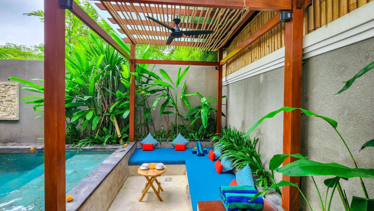 Tropical Balinese outdoor lounge with blue cushions on wooden deck, bamboo pergola, ceiling fan, lush greenery, and turquoise plunge pool. - Bali Villas