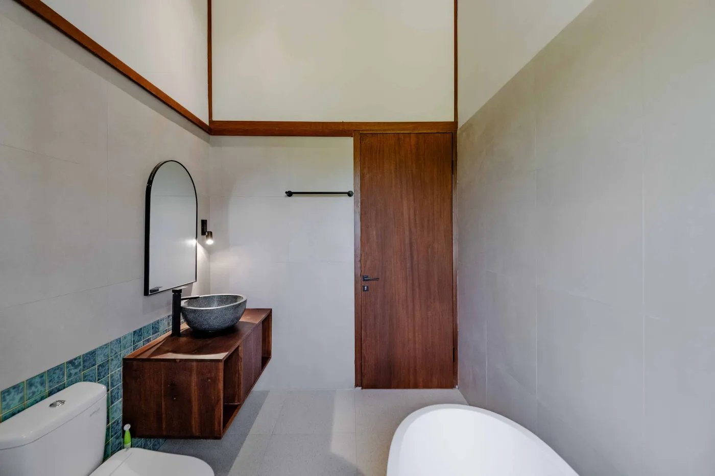 Modern bathroom with white freestanding tub, glass-enclosed teal-tiled shower, white toilet, and wooden shutters overlooking greenery. - Bali Villas