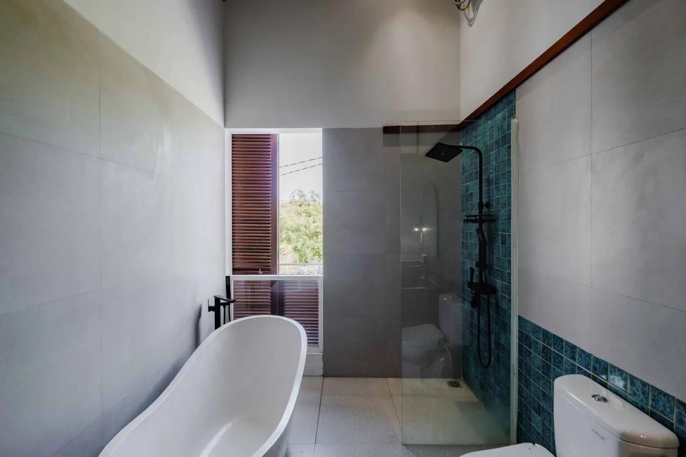 Modern bathroom with white freestanding tub, glass-enclosed teal-tiled shower, white toilet, and wooden shutters overlooking greenery. - Bali Villas