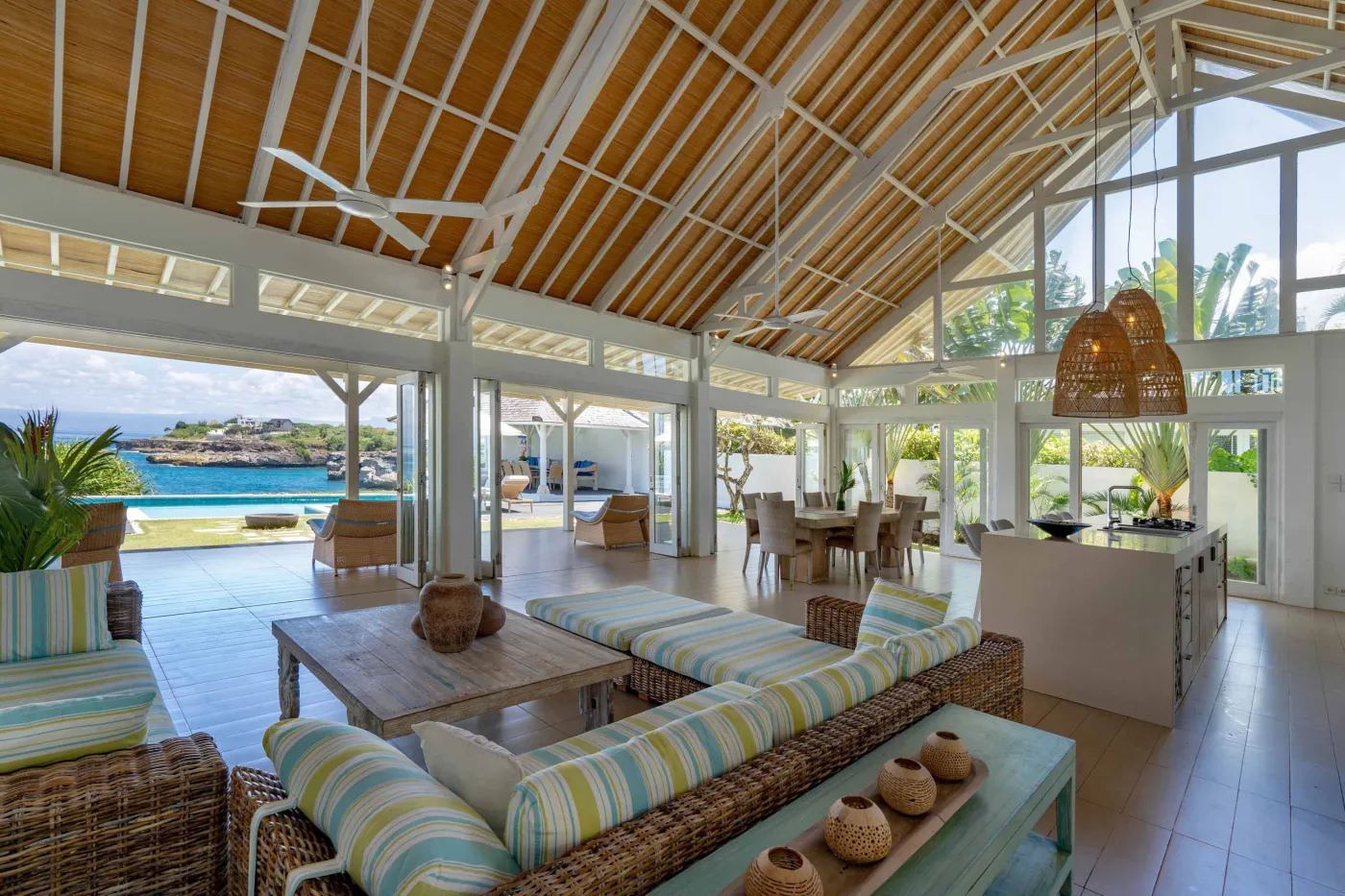 Spacious modern Balinese-style kitchen with high wooden ceiling, rattan pendant lights, white island bar, fresh fruits, and tropical garden views through large windows. - Bali Villas