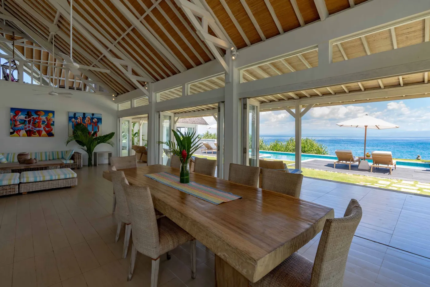 Spacious modern Balinese-style kitchen with high wooden ceiling, rattan pendant lights, white island bar, fresh fruits, and tropical garden views through large windows. - Bali Villas