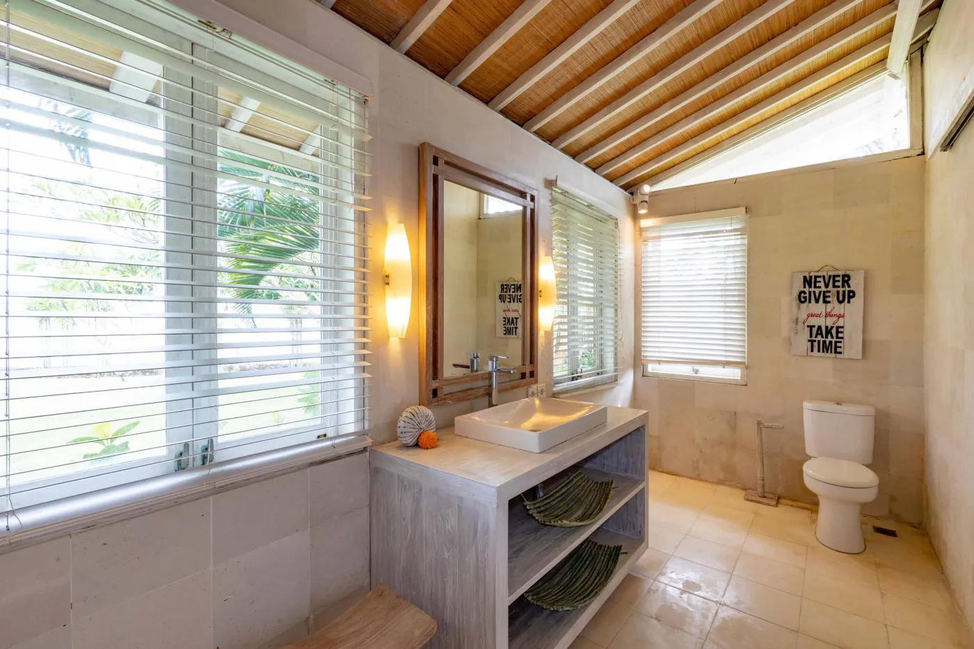 Spacious modern Balinese-style kitchen with high wooden ceiling, rattan pendant lights, white island bar, fresh fruits, and tropical garden views through large windows. - Bali Villas