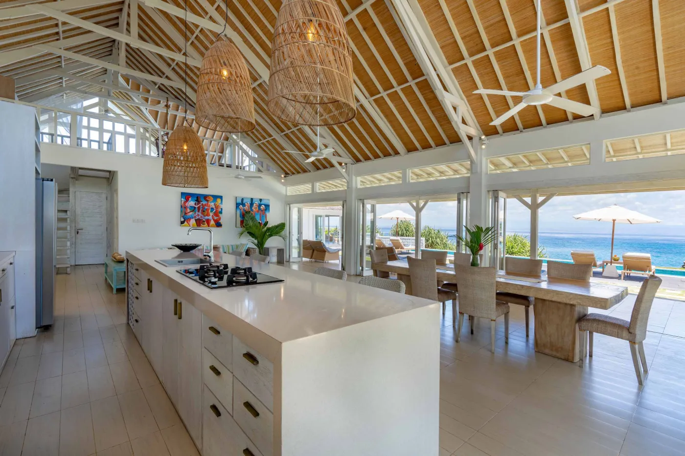 Spacious modern Balinese-style kitchen with high wooden ceiling, rattan pendant lights, white island bar, fresh fruits, and tropical garden views through large windows. - Bali Villas