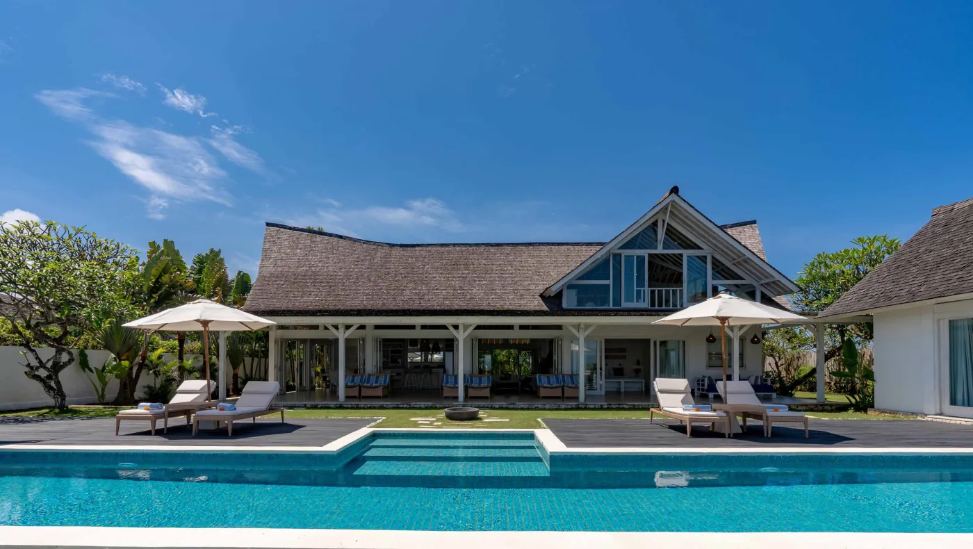 Spacious modern Balinese-style kitchen with high wooden ceiling, rattan pendant lights, white island bar, fresh fruits, and tropical garden views through large windows. - Bali Villas