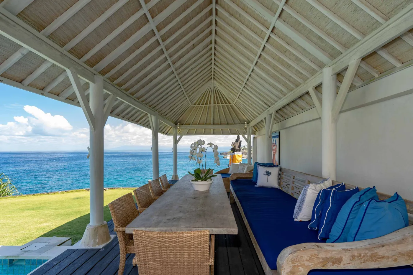 Spacious modern Balinese-style kitchen with high wooden ceiling, rattan pendant lights, white island bar, fresh fruits, and tropical garden views through large windows. - Bali Villas