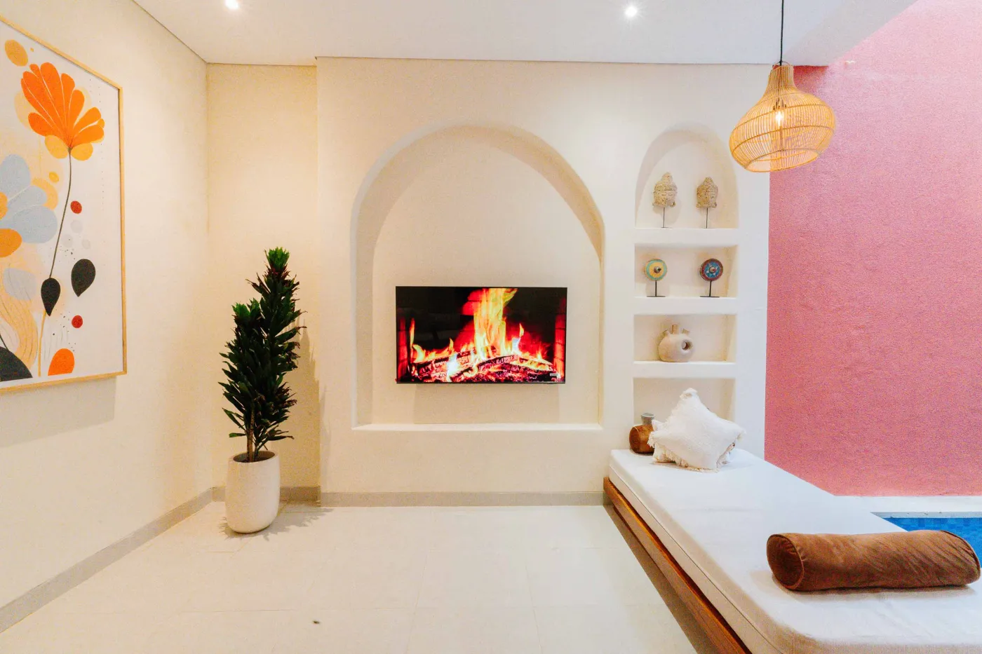 Cozy modern bedroom interior with wooden walls, low white bed, fireplace TV, potted plants, abstract art, and tropical fruit bowl on woven stool. - Bali Villas