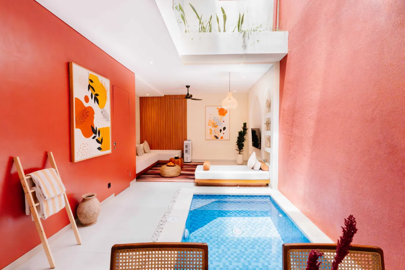 Cozy modern bedroom interior with wooden walls, low white bed, fireplace TV, potted plants, abstract art, and tropical fruit bowl on woven stool. - Bali Villas