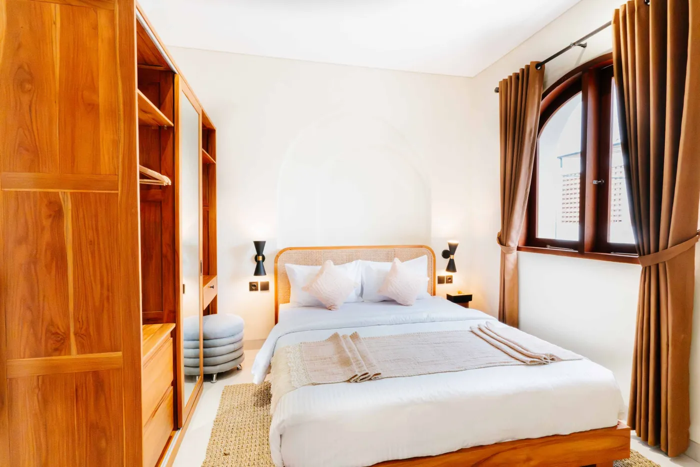 Cozy modern bedroom interior with wooden walls, low white bed, fireplace TV, potted plants, abstract art, and tropical fruit bowl on woven stool. - Bali Villas