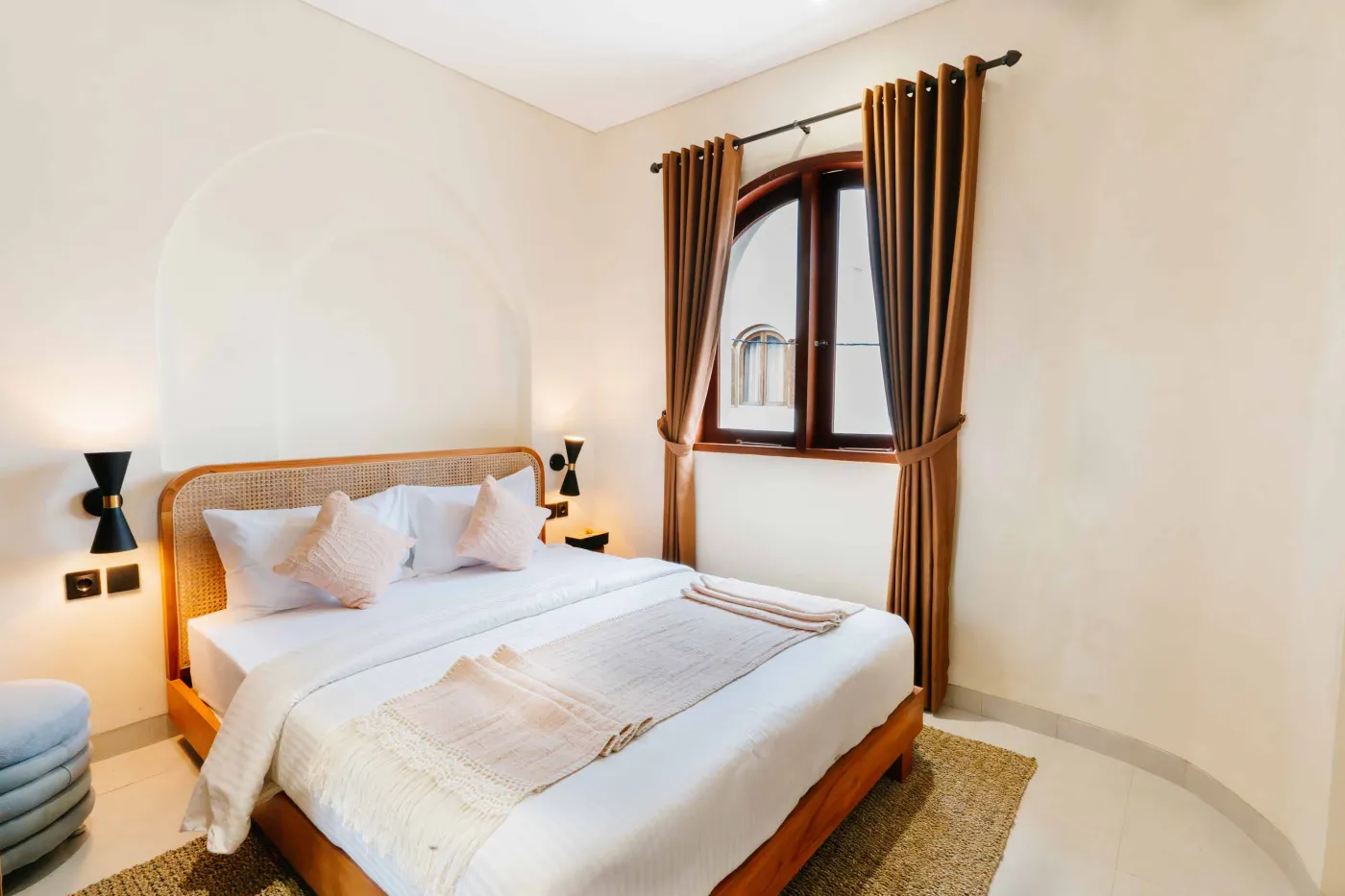 Cozy modern bedroom interior with wooden walls, low white bed, fireplace TV, potted plants, abstract art, and tropical fruit bowl on woven stool. - Bali Villas