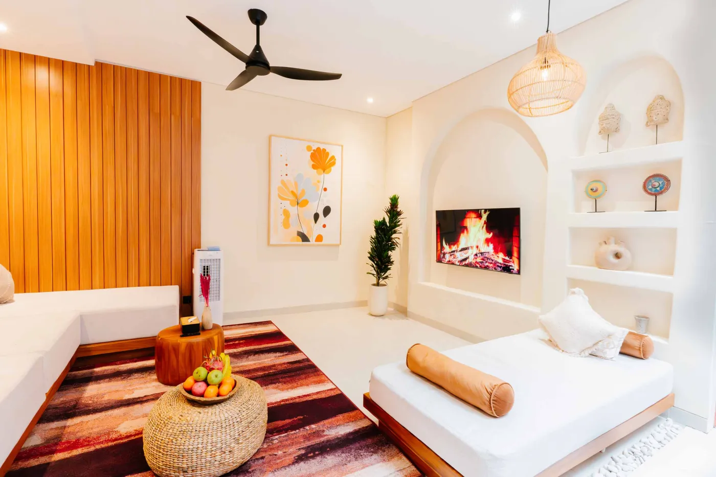 Compact white-tiled bathroom with round black mirror, woven pendant light, frangipani flowers on shelf, white sink, toilet, and folded towels. - Bali Villas