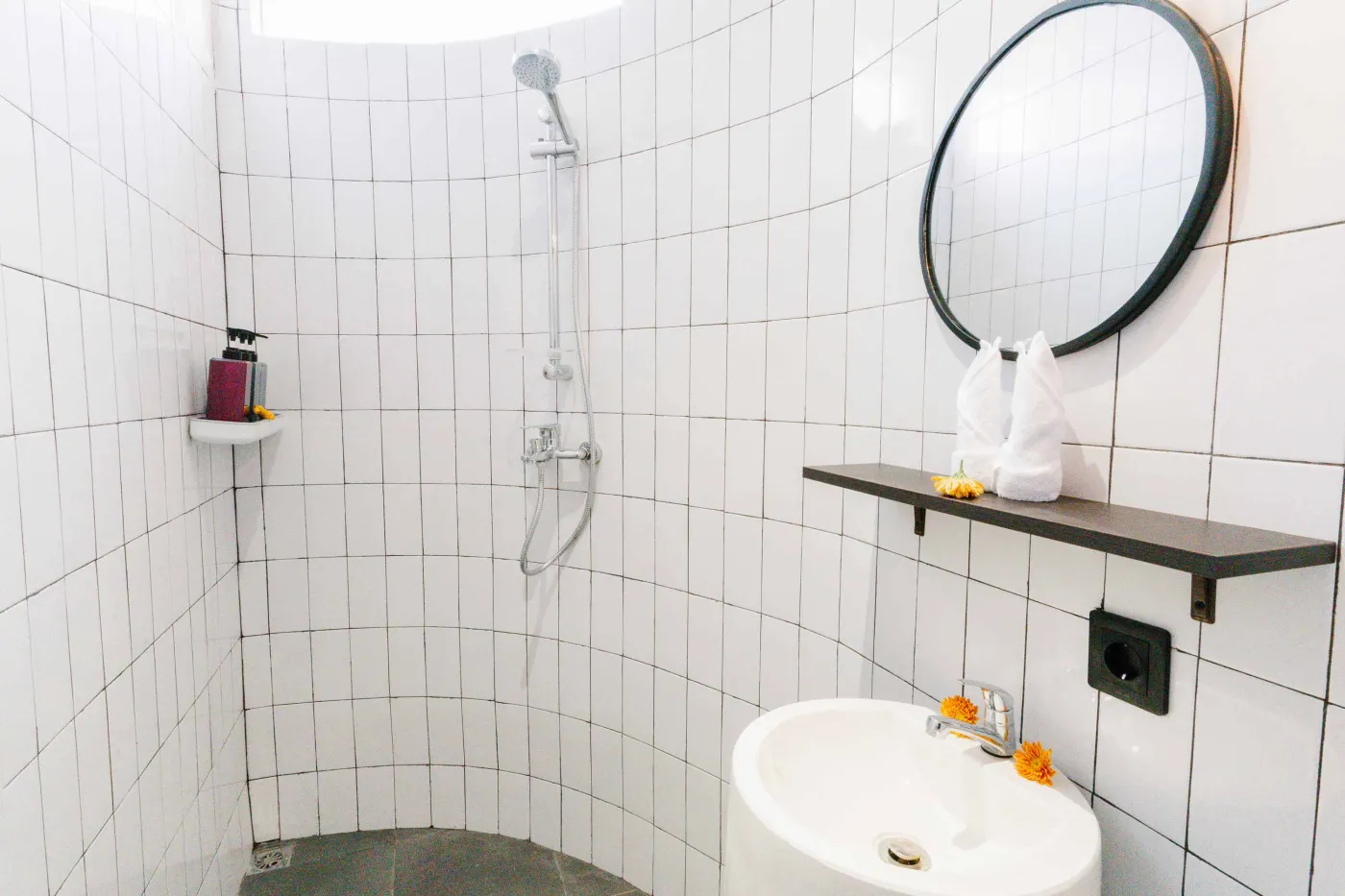 Compact white-tiled bathroom with round black mirror, woven pendant light, frangipani flowers on shelf, white sink, toilet, and folded towels. - Bali Villas