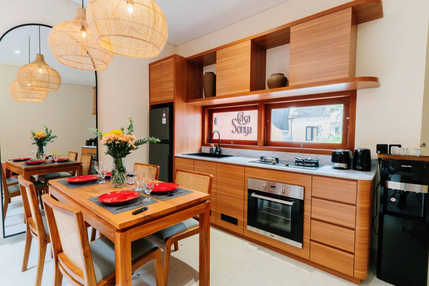 Compact white-tiled bathroom with round black mirror, woven pendant light, frangipani flowers on shelf, white sink, toilet, and folded towels. - Bali Villas
