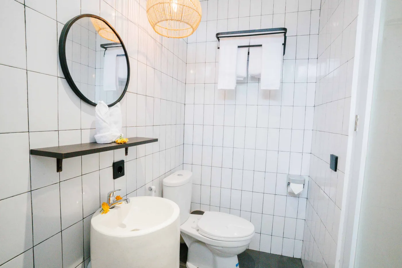 Compact white-tiled bathroom with round black mirror, woven pendant light, frangipani flowers on shelf, white sink, toilet, and folded towels. - Bali Villas