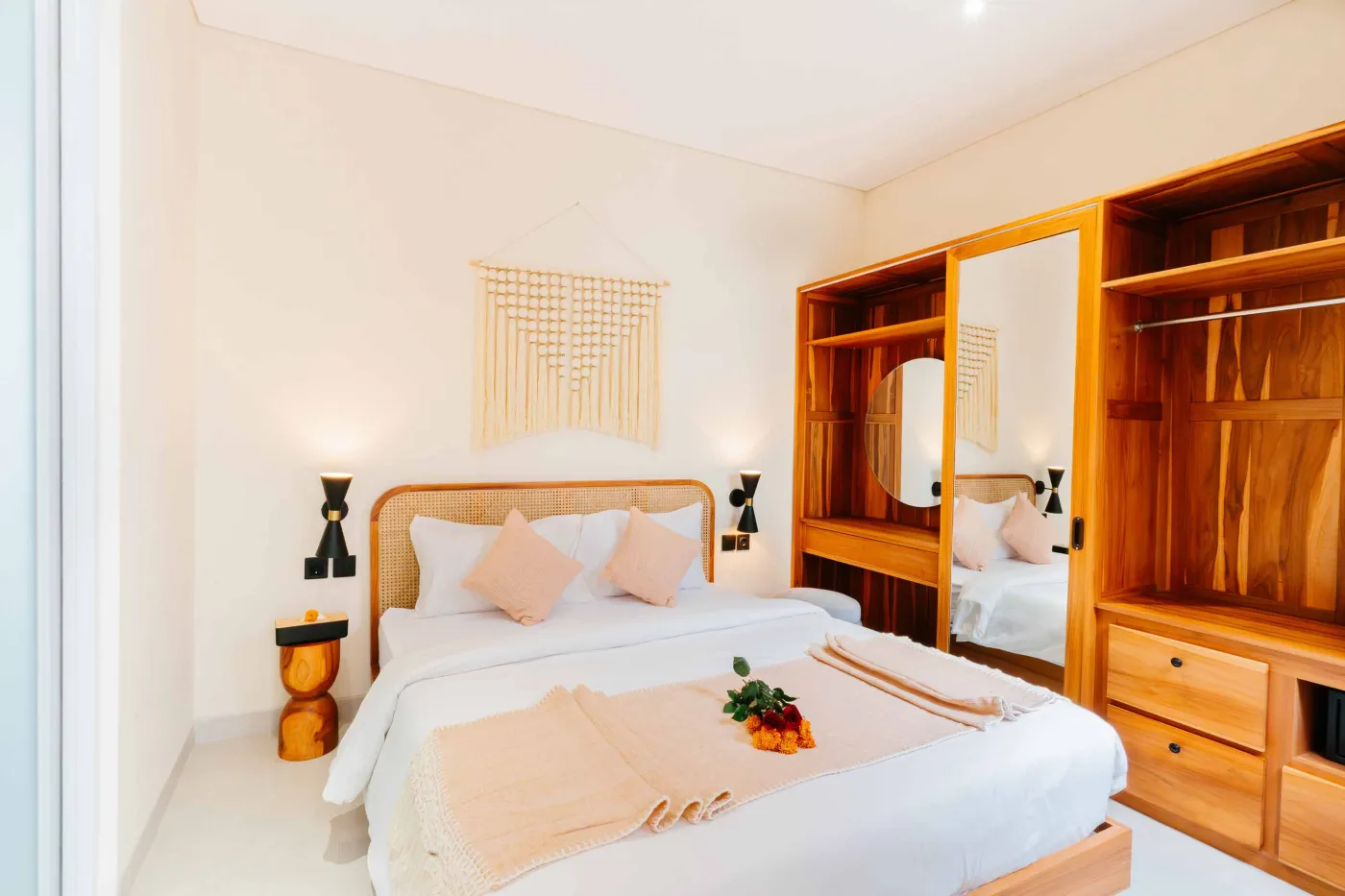 Compact white-tiled bathroom with round black mirror, woven pendant light, frangipani flowers on shelf, white sink, toilet, and folded towels. - Bali Villas