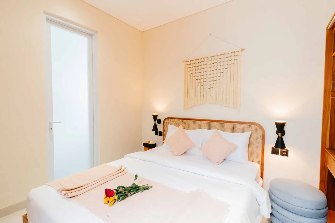 Compact white-tiled bathroom with round black mirror, woven pendant light, frangipani flowers on shelf, white sink, toilet, and folded towels. - Bali Villas