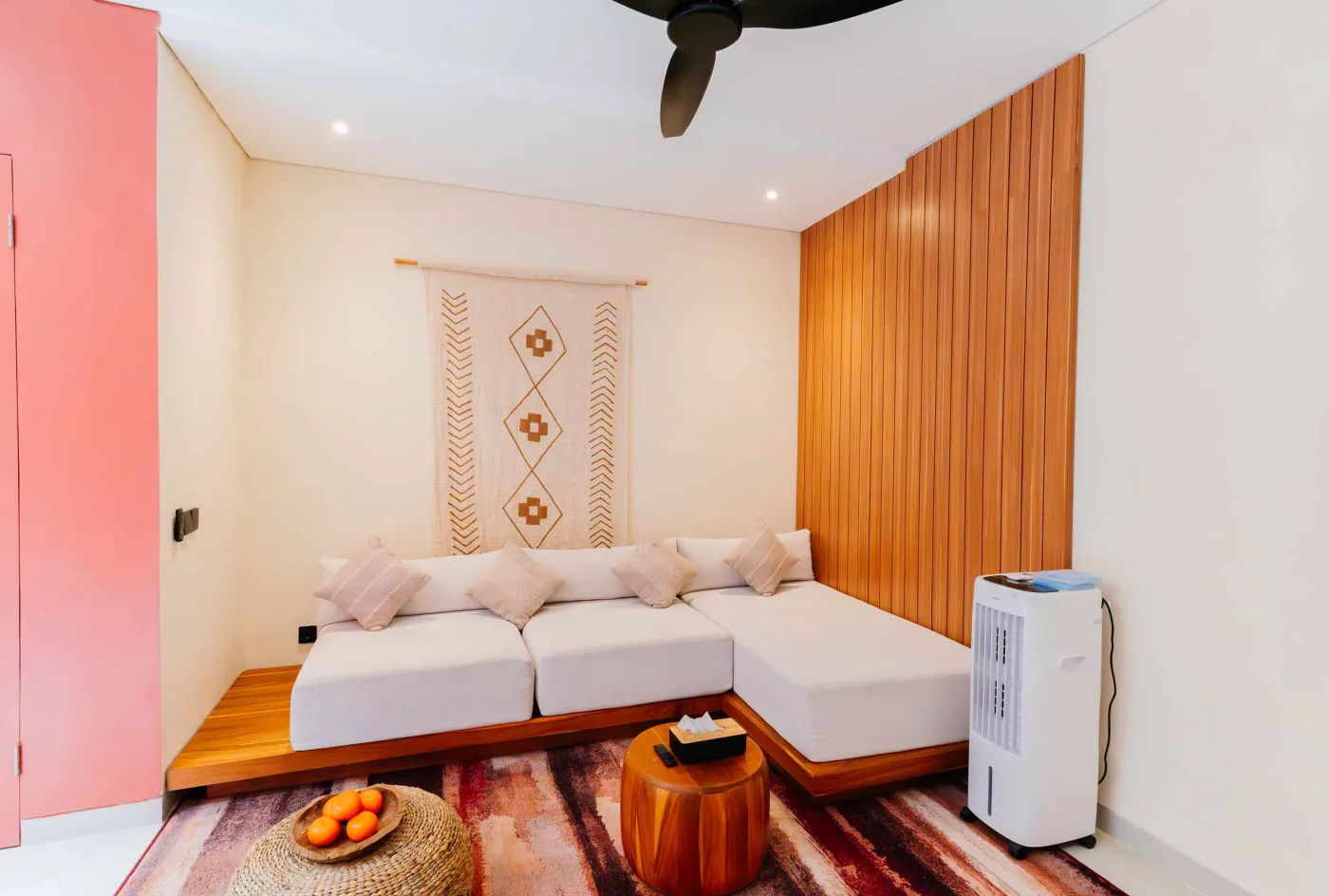 Compact white-tiled bathroom with round black mirror, woven pendant light, frangipani flowers on shelf, white sink, toilet, and folded towels. - Bali Villas